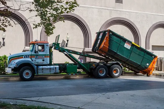 Garbage truck unloading a large green dumpster in front of a building