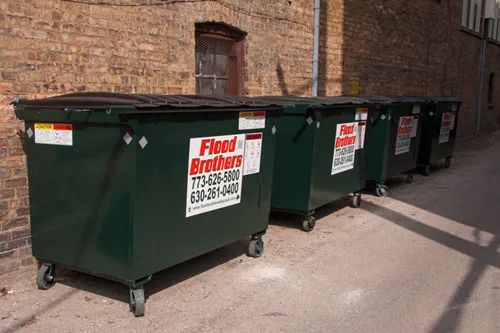 Row of green commercial dumpsters with Flood Brothers branding beside a brick building